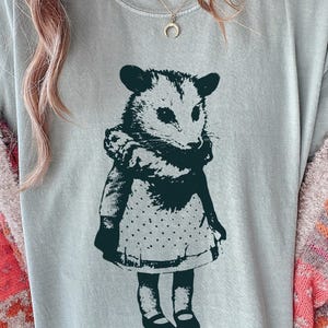 Pode incluir: Camiseta azul claro com uma ilustra&ccedil;&atilde;o verde escuro de um gamb&aacute; usando um vestido com uma saia de bolinhas. O gamb&aacute; est&aacute; em p&eacute;. A camisa tem gola redonda.