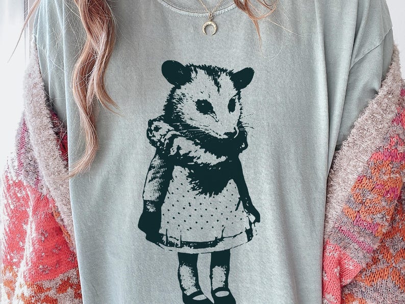Pode incluir: Camiseta azul claro com uma ilustra&ccedil;&atilde;o verde escuro de um gamb&aacute; usando um vestido com uma saia de bolinhas. O gamb&aacute; est&aacute; em p&eacute;. A camisa tem gola redonda.