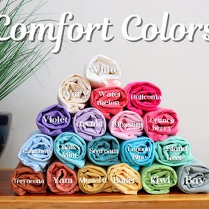 May include: A pyramid of rolled-up t-shirts in various colors, including white, violet, and seafoam. The text "Comfort Colors" is displayed at the top. Each shirt is labeled with its color name, such as "Watermelon" and "Mustard".