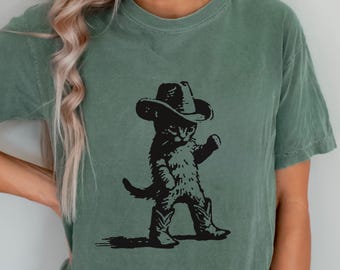 May include: Sage green t-shirt featuring a black graphic of a cat wearing a cowboy hat and boots. The cat is depicted in a walking pose, with one arm raised. The shirt has a relaxed fit and short sleeves.