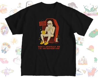May include: Black t-shirt featuring a cartoon mummy in a coffin, holding a sign that reads "Do Not Disturb Seriously." The mummy is wearing bunny slippers and the text "Don't Unwrap Me I'm Introverting" is below the image.