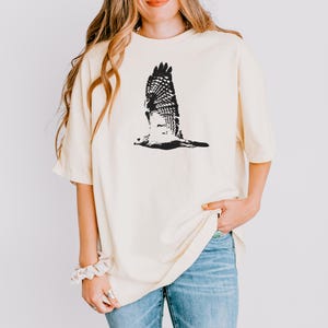May include: Cream-colored t-shirt featuring a black graphic of a hawk in flight. The bird's wings are spread wide, showcasing detailed feather patterns. The shirt has short sleeves and a relaxed fit, paired with blue jeans.