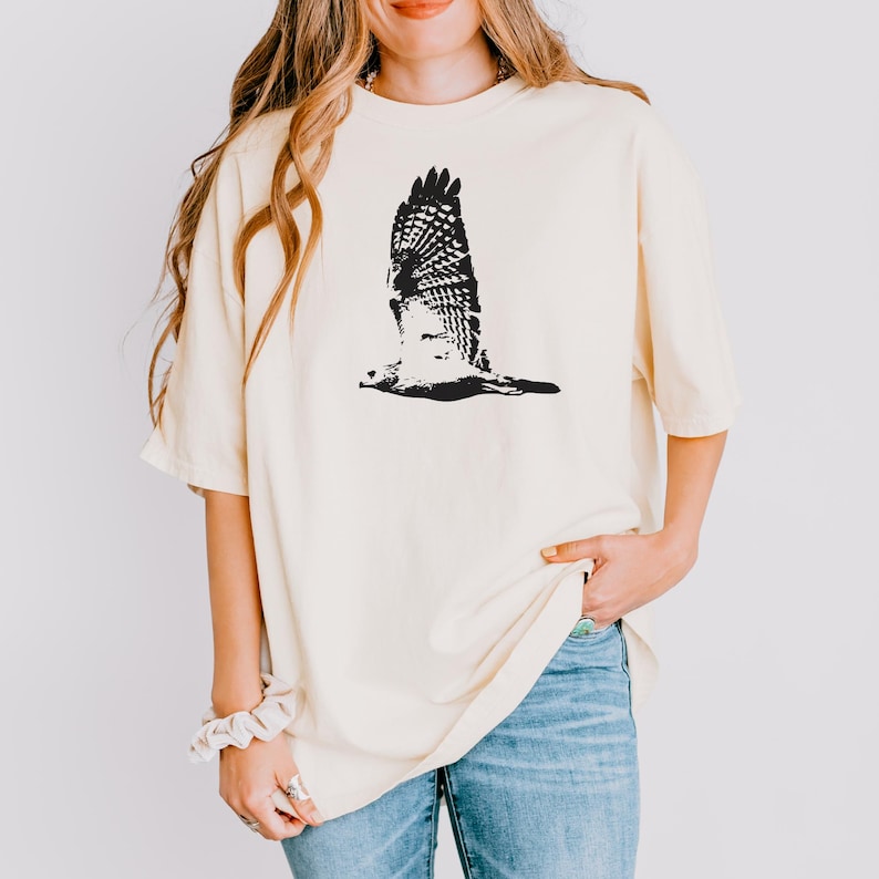 May include: Cream-colored t-shirt featuring a black graphic of a hawk in flight. The bird's wings are spread wide, showcasing detailed feather patterns. The shirt has short sleeves and a relaxed fit, paired with blue jeans.