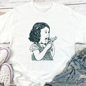 Puede incluir: Camiseta blanca con una ilustraci&oacute;n en blanco y negro de una ni&ntilde;a comiendo una babosa. El gr&aacute;fico es un dibujo lineal detallado, con la expresi&oacute;n de sorpresa de la ni&ntilde;a. La camiseta est&aacute; hecha de un material suave y c&oacute;modo.
