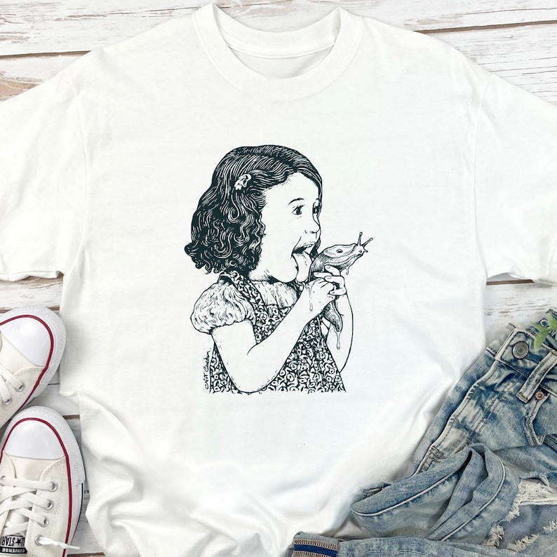 Puede incluir: Camiseta blanca con una ilustraci&oacute;n en blanco y negro de una ni&ntilde;a comiendo una babosa. El gr&aacute;fico es un dibujo lineal detallado, con la expresi&oacute;n de sorpresa de la ni&ntilde;a. La camiseta est&aacute; hecha de un material suave y c&oacute;modo.