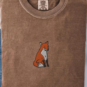 Peut inclure: T-shirt Comfort Colors marron avec un motif de renard brodé. Le renard est orange, blanc et noir. Le t-shirt a un col rond et l'étiquette indique XL-TG-EG.