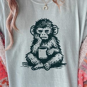 May include: A sage green short-sleeve t-shirt featuring a dark green graphic print of a monkey sitting with its hand on its chin while holding a coffee mug. The monkey illustration has a textured, woodcut-style appearance on the front of the shirt.