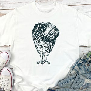 Puede incluir: Camiseta blanca con una ilustraci&oacute;n en blanco y negro de un b&uacute;ho inclinado. Los ojos del b&uacute;ho son grandes y redondos, y sus plumas est&aacute;n detalladas. La camiseta se combina con vaqueros y zapatillas blancas.