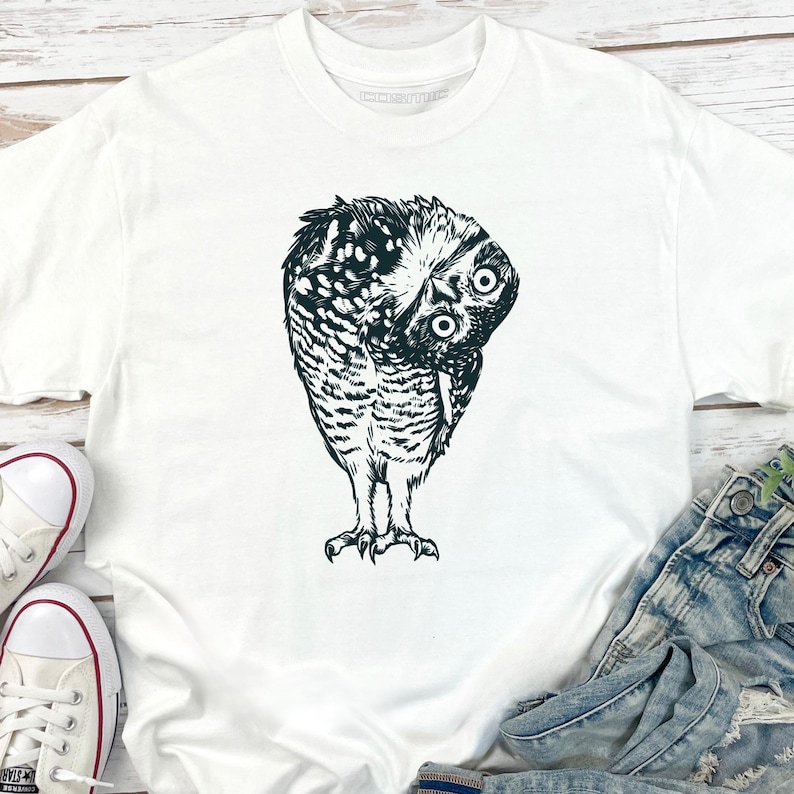 Puede incluir: Camiseta blanca con una ilustraci&oacute;n en blanco y negro de un b&uacute;ho inclinado. Los ojos del b&uacute;ho son grandes y redondos, y sus plumas est&aacute;n detalladas. La camiseta se combina con vaqueros y zapatillas blancas.