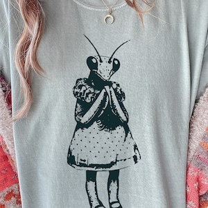 Op de afbeelding: Lichtblauw T-shirt met een donkergroene illustratie van een bidsprinkhaan in een jurk. De bidsprinkhaan heeft de handen gevouwen. De jurk heeft een stippenpatroon.