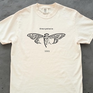 May include: Cream-coloured t-shirt featuring a black cicada design and the word "Everywhere" above it. The number "3301" is printed below the insect. The shirt is made of a soft, comfortable material.