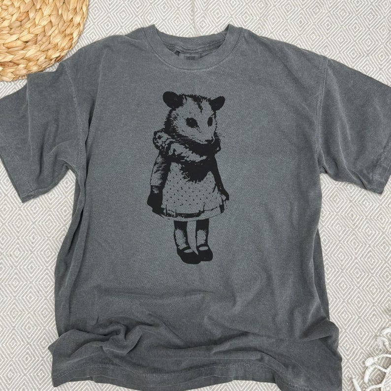 Pode incluir: Camiseta cinza com um gr&aacute;fico preto de um gamb&aacute; com um vestido. O gamb&aacute; &eacute; retratado em p&eacute;, com os bra&ccedil;os ao lado. A camiseta tem gola redonda e mangas curtas. O tecido da camiseta parece macio e confort&aacute;vel.