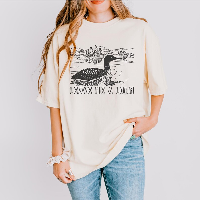 May include: Cream-colored t-shirt featuring a black and white graphic of a loon swimming on a lake, with mountains and trees in the background. The text "LEAVE ME A LOON" is printed below the image. The shirt is a relaxed fit.