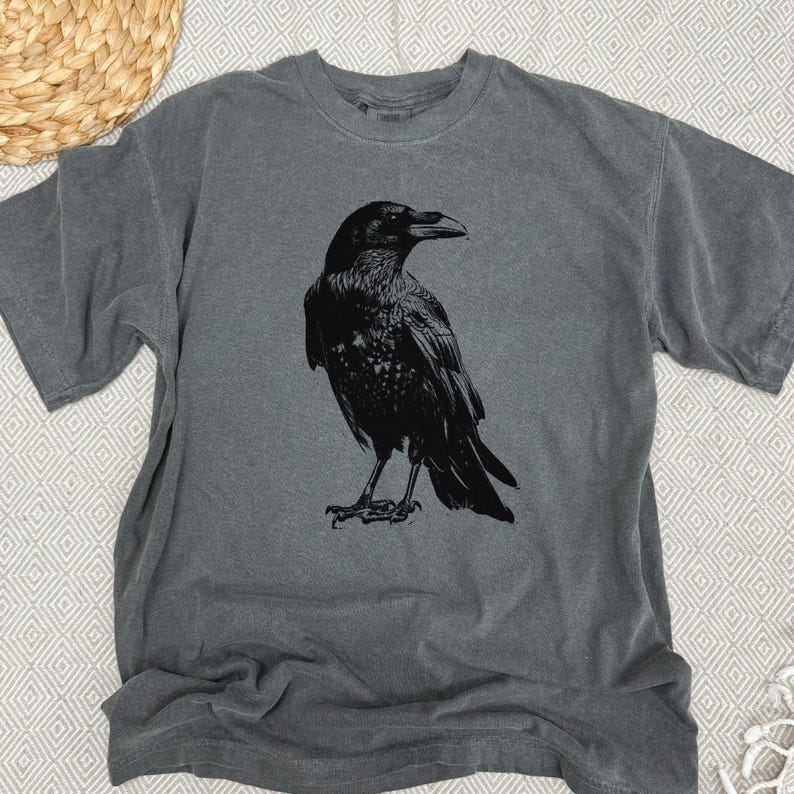 May include: A gray t-shirt featuring a detailed black illustration of a raven. The bird is depicted in a standing pose, with its wings slightly folded. The shirt has a classic crew neck and short sleeves.