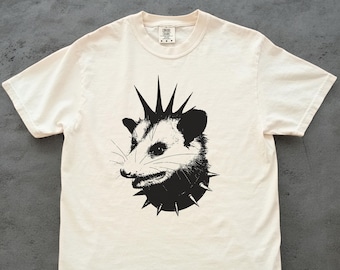 Könnte beinhalten: Cremefarbenes T-Shirt mit einer schwarz-weißen Grafik eines Opossums mit Irokesenschnitt und Stachelhalsband. Das Opossum blickt nach rechts. Das Shirt hat einen Rundhalsausschnitt und kurze Ärmel.