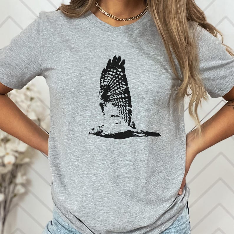May include: A light gray t-shirt featuring a black graphic of a bird in flight. The bird's wings are spread wide, showcasing detailed feather patterns. The shirt is a crew neck style.
