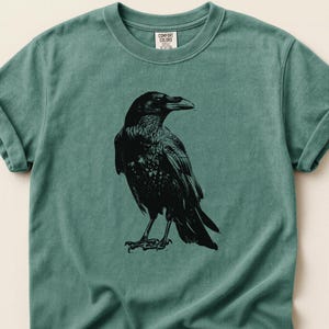 May include: A teal-colored t-shirt featuring a detailed black illustration of a raven. The raven is depicted in a side profile, showcasing its beak and feathers. The shirt has rolled-up sleeves and a Comfort Colors tag.