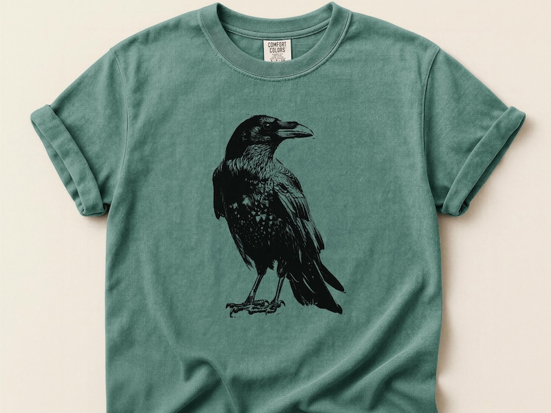 May include: A teal-colored t-shirt featuring a detailed black illustration of a raven. The raven is depicted in a side profile, showcasing its beak and feathers. The shirt has rolled-up sleeves and a Comfort Colors tag.