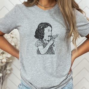 Puede incluir: Camiseta gris con una ilustraci&oacute;n en blanco y negro de una ni&ntilde;a comiendo un caracol. El gr&aacute;fico es un dibujo de estilo vintage. La camiseta tiene mangas cortas y cuello redondo.