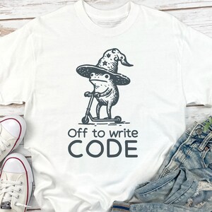 May include: White t-shirt with a black graphic of a frog wearing a wizard hat and riding a scooter. The text "Off to write CODE" is below the graphic. The shirt is paired with blue jeans and white sneakers.
