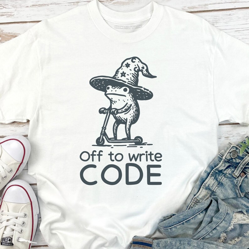 May include: White t-shirt with a black graphic of a frog wearing a wizard hat and riding a scooter. The text "Off to write CODE" is below the graphic. The shirt is paired with blue jeans and white sneakers.