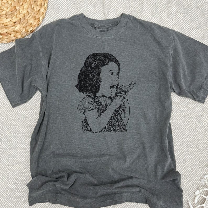 Puede incluir: Camiseta gris con una ilustraci&oacute;n en blanco y negro de una ni&ntilde;a comiendo una babosa. La camiseta gr&aacute;fica tiene mangas cortas y cuello redondo. La camiseta est&aacute; hecha de un material suave y c&oacute;modo.