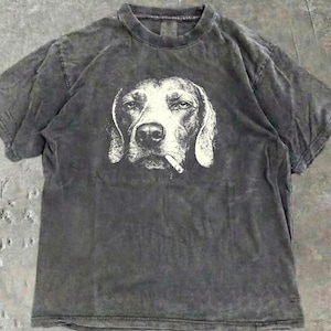 Puede incluir: Camiseta gris carbón con un gráfico blanco de la cara de un perro con un cigarrillo en la boca. La camiseta tiene cuello redondo y mangas cortas. La tela tiene un aspecto descolorido y vintage.