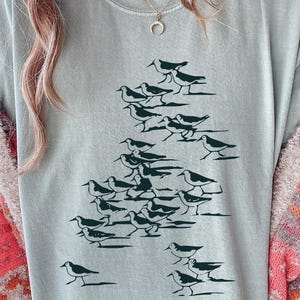 May include: A light blue t-shirt featuring a flock of birds in a dark green color. The birds are arranged in a triangular formation. The shirt is made of a soft material.