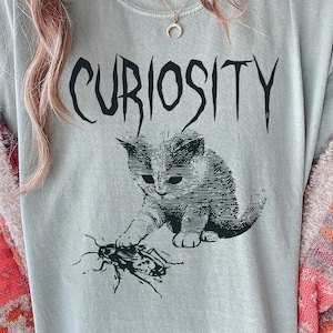 Puede incluir: Camiseta gris claro con la palabra "CURIOSITY" en fuente gótica negra. Una ilustración en blanco y negro de un gatito jugando con una cucaracha está debajo del texto. La camiseta es de estilo casual.