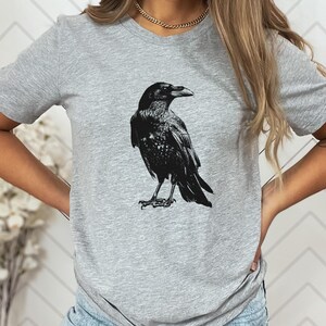 May include: A gray t-shirt featuring a detailed black illustration of a raven. The raven is depicted in a standing pose, with its wings slightly spread. The t-shirt is made of a soft, comfortable material.