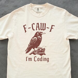 May include: Off-white t-shirt featuring a brown raven illustration above a typewriter, with the text "F-CAW-F" and "I'm Coding". The shirt has a crew neck and short sleeves, made of a soft, light fabric.