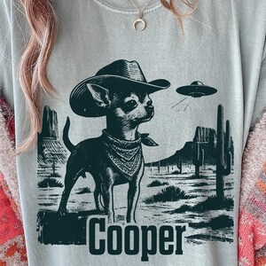 May include: A light grey t-shirt with a black and white illustration of a Chihuahua wearing a cowboy hat and bandana, with a UFO in the background. The name "Cooper" is printed below the dog in a bold font.
