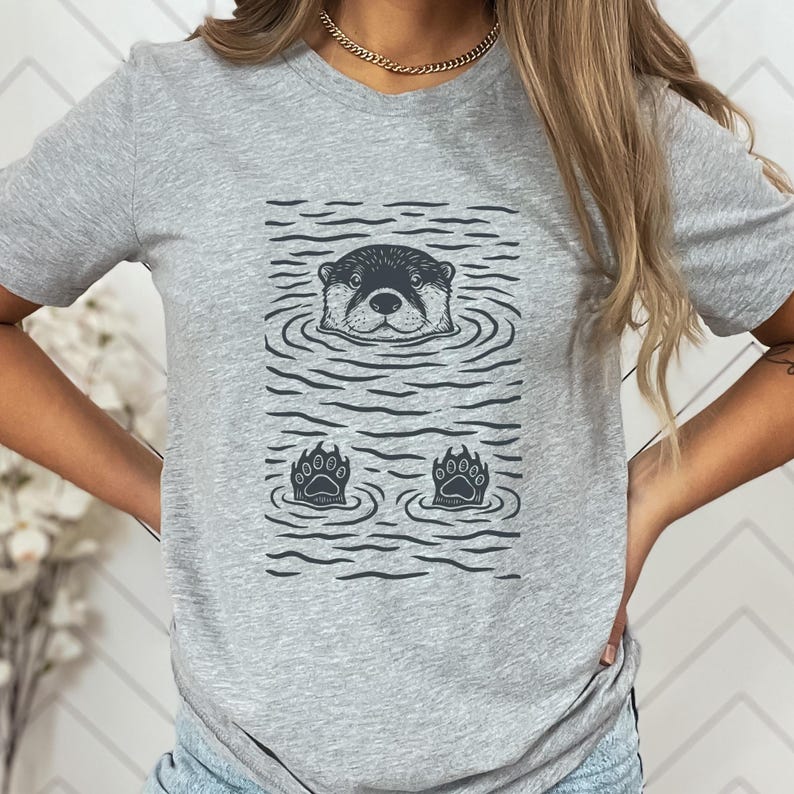 May include: Gray t-shirt featuring a black and white illustration of an otter in water. The design includes the otter's head, two paws, and water ripples. The shirt is made of a soft material.