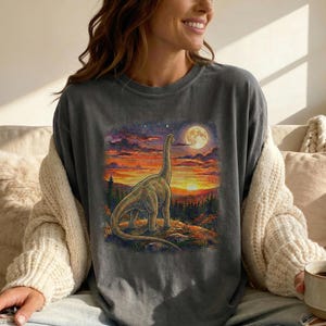 May include: Dark grey t-shirt with a Brachiosaurus dinosaur illustration set against a sunset and full moon. The design features orange, yellow, and purple hues. The shirt has a relaxed fit.