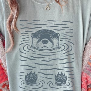May include: A light blue t-shirt features a graphic of an otter in water, with its head and paws visible. The otter is surrounded by water ripples. The shirt is a comfortable, casual style.