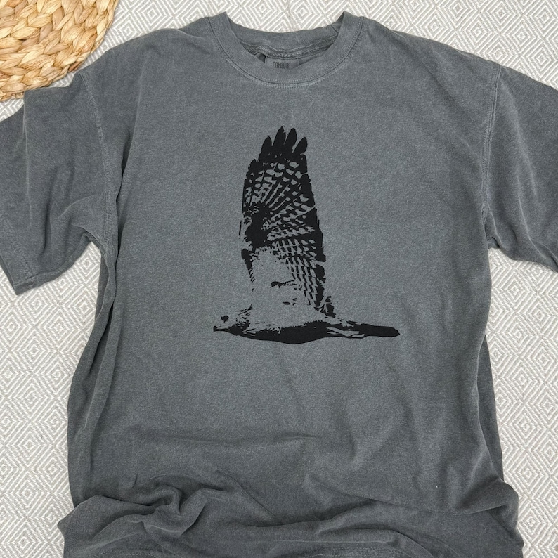 May include: A gray short-sleeved t-shirt featuring a black graphic of a hawk in flight. The bird's wings are spread wide, showcasing detailed feather patterns. The shirt has a crew neck and a relaxed fit, suitable for casual wear.