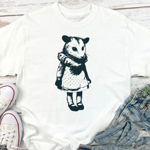 Pode incluir: Camiseta branca com uma ilustra&ccedil;&atilde;o em preto e branco de um gamb&aacute; com um vestido. O gamb&aacute; &eacute; retratado em um estilo vintage, com uma gola com babados e um vestido de bolinhas. A camiseta &eacute; mostrada com jeans e t&ecirc;nis.