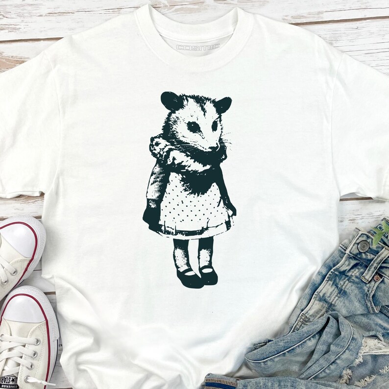 Pode incluir: Camiseta branca com uma ilustra&ccedil;&atilde;o em preto e branco de um gamb&aacute; com um vestido. O gamb&aacute; &eacute; retratado em um estilo vintage, com uma gola com babados e um vestido de bolinhas. A camiseta &eacute; mostrada com jeans e t&ecirc;nis.