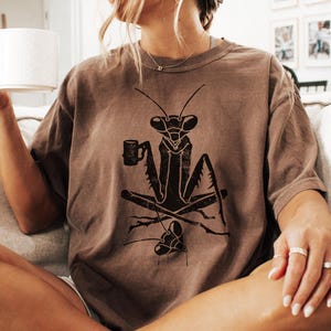 May include: Brown t-shirt featuring a black graphic of a praying mantis holding a mug. The insect is depicted with crossed legs and a small design below. The shirt has a relaxed fit and short sleeves.