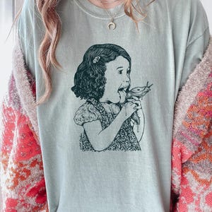 Puede incluir: Camiseta verde salvia con una ilustraci&oacute;n en blanco y negro de una ni&ntilde;a comiendo una babosa. El gr&aacute;fico es un dibujo de estilo vintage con sombreado detallado. La expresi&oacute;n de la ni&ntilde;a es de sorpresa.