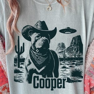 Pode incluir: Camiseta cinza claro com um gráfico preto de um cachorro usando um chapéu de cowboy e bandana, com uma paisagem desértica e um OVNI. O nome "Cooper" é impresso abaixo.