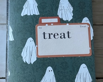 May include: A dark green notebook with a repeating pattern of white ghost illustrations. The word "treat" is displayed in a rectangular label. The notebook is a Halloween-themed item.