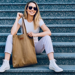May include: A woman wearing sunglasses, a white t-shirt, and light purple pants sits on gray steps, holding a large brown tote bag. She is also wearing white high-top sneakers. The tote bag has a brown leather patch.