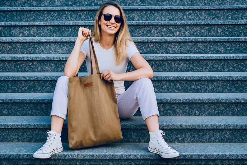 May include: A woman wearing sunglasses, a white t-shirt, and light purple pants sits on gray steps, holding a large brown tote bag. She is also wearing white high-top sneakers. The tote bag has a brown leather patch.
