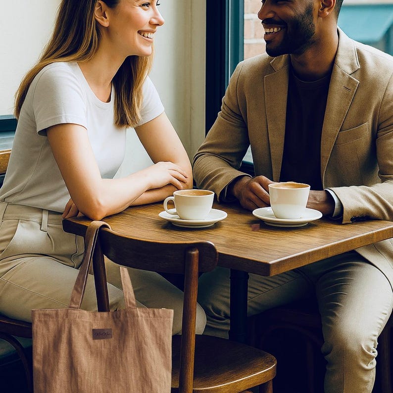 May include: A couple smiles while sitting at a wooden table in a cafe. Two white coffee cups and saucers sit on the table. A tan tote bag hangs from a wooden chair. The man wears a tan blazer, and the woman wears a white t-shirt.