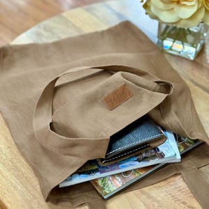 May include: A tan tote bag with long handles, filled with a wallet and magazines. The bag has a small leather patch with a logo. The bag is sitting on a wooden table.