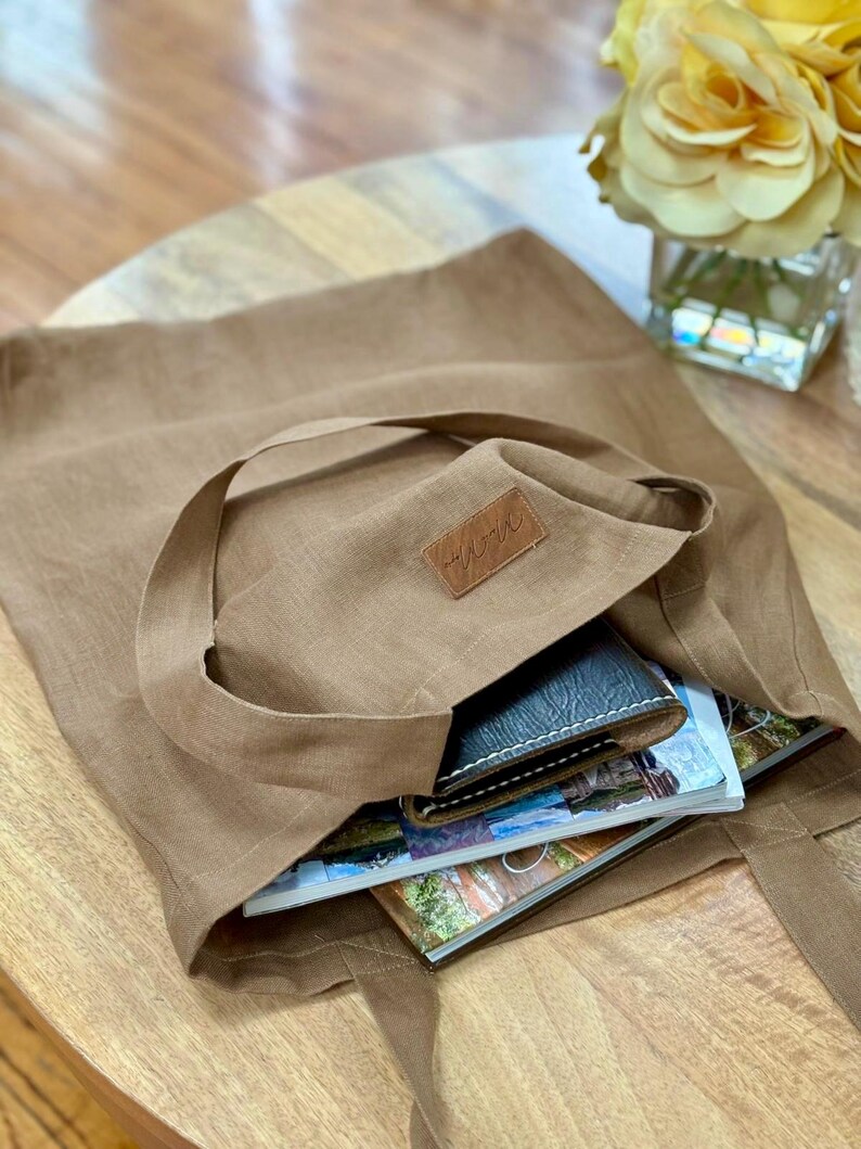 May include: A tan tote bag with long handles, filled with a wallet and magazines. The bag has a small leather patch with a logo. The bag is sitting on a wooden table.