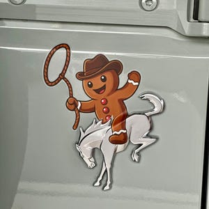 May include: A whimsical sticker featuring a gingerbread man cowboy riding a white horse. The gingerbread man wears a brown cowboy hat and holds a lasso. The sticker is affixed to a light gray surface.
