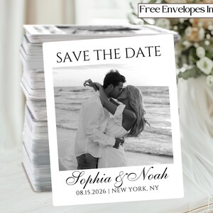 May include: A stack of white "Save the Date" cards featuring a black and white photo of a couple kissing on a beach. The cards include the names "Sophia & Noah" and the date "08.15.2026 | NEW YORK, NY". Text at the top reads "Free Envelopes Included".