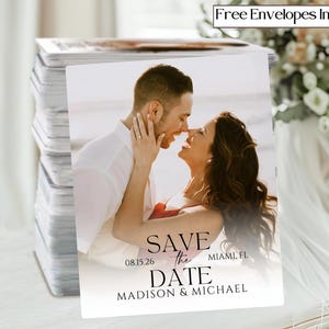 May include: A stack of save-the-date cards featuring a couple embracing on a beach. The cards have the text "Save the Date" with the date 08.15.26 and the names Madison & Michael, Miami, FL. The words "Free Envelopes Included" are in the top right corner.